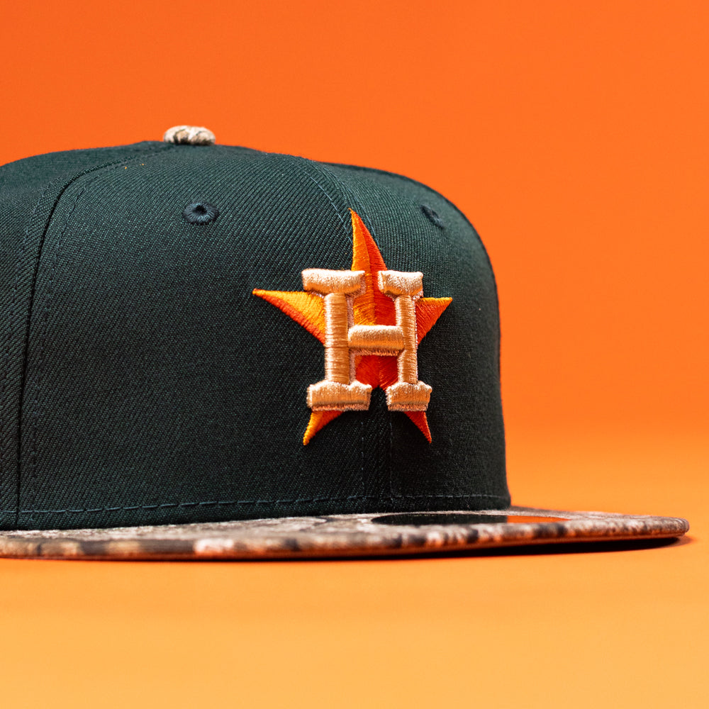 The Greenhand RTC Oil Houston Astros New Era Hat 5950 Fitted features a dark green color, a camouflage brim, and an embroidered orange and gold H with a star on the front, shown against an orange background.