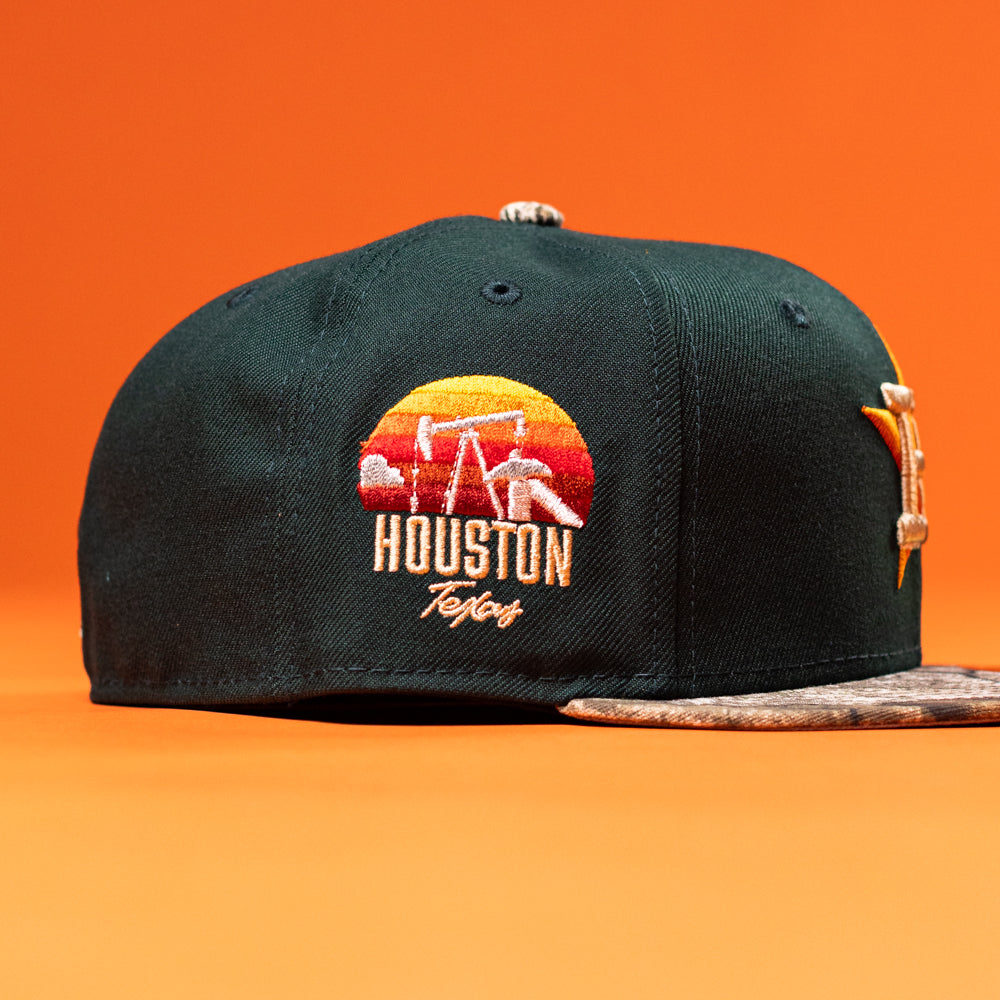 The Greenhand RTC Oil Houston Astros New Era Hat 5950 Fitted is a black New Era hat with a sunset patch of oil pumps, "Houston Texas" side embroidery, and a brown textured visor—ideal for Astros fans. Background is solid orange.