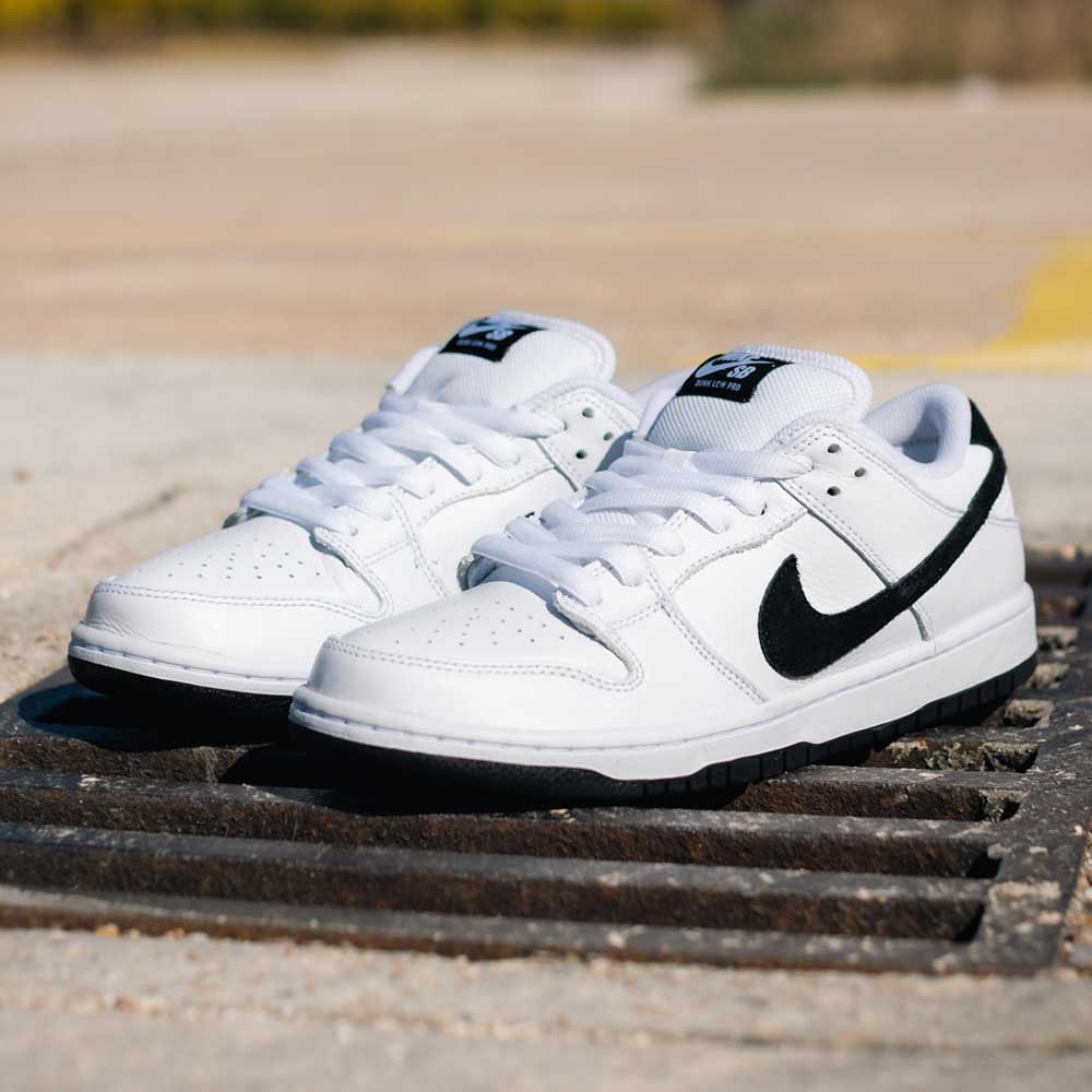 The HF3704100 White Black Nike SB Dunk Low Pro Suede Leather Shoe is displayed outdoors on a metal grate in bright sunlight, highlighting its classic black swooshes, black soles, and stylish skateboarding design.