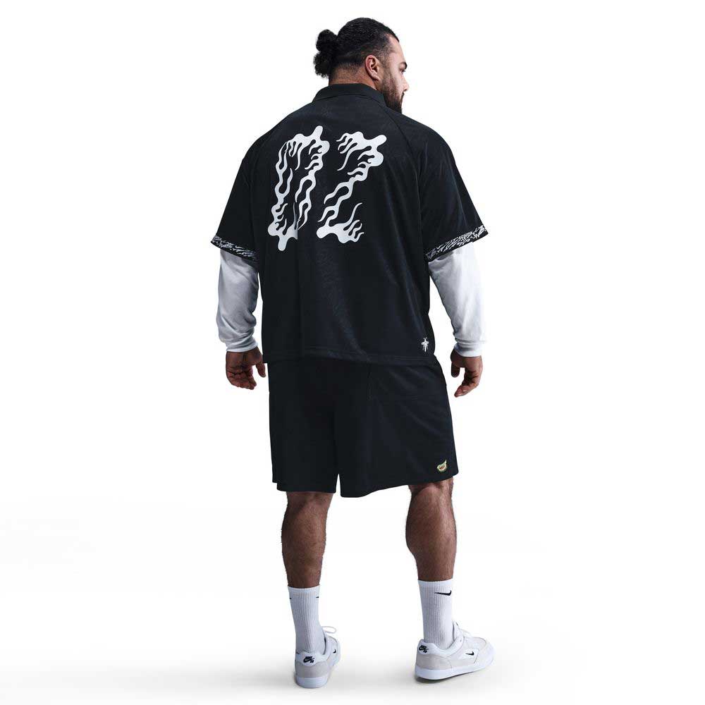 A man faces away wearing the HJ 2965010 Black Nike SB Dri-FIT Jacquard Skate Jersey with white back graphics, paired with black shorts, a white long-sleeve shirt underneath, white socks, and white sneakers, set against a plain white background.