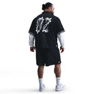 A man faces away wearing the HJ 2965010 Black Nike SB Dri-FIT Jacquard Skate Jersey with white back graphics, paired with black shorts, a white long-sleeve shirt underneath, white socks, and white sneakers, set against a plain white background.