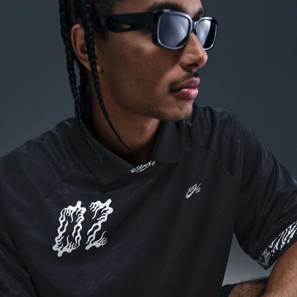A man with braided hair and dark sunglasses wears the HJ 2965010 Black Nike SB Dri-FIT Jacquard Skateboarding Jersey, which features a bold wavy 02 graphic. The dark background enhances the stylish, moody vibe.