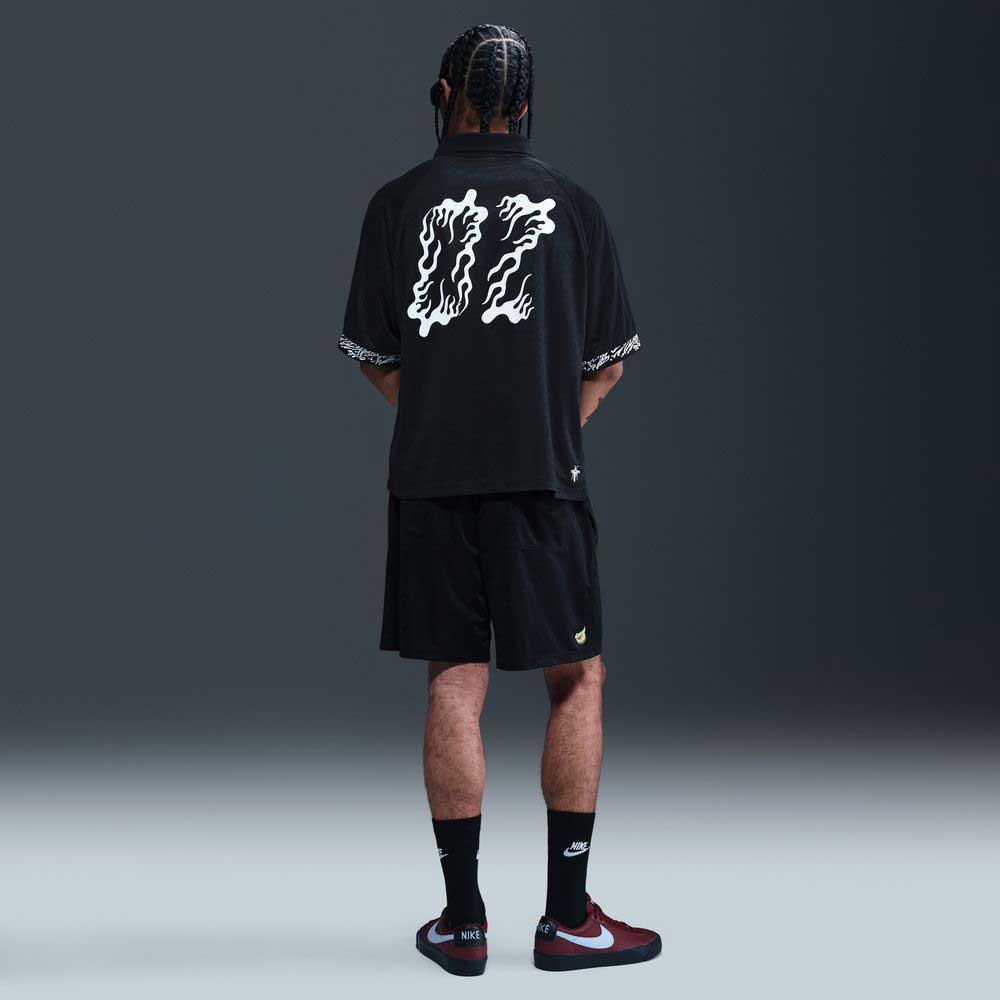 A person with braided hair faces away, wearing the HJ 2965010 Black Nike SB Dri-FIT Jacquard Skate Jersey Skateboarding, black shorts, black Nike socks, and maroon and white Nike sneakers against a dark background.