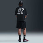A person with braided hair faces away, wearing the HJ 2965010 Black Nike SB Dri-FIT Jacquard Skate Jersey Skateboarding, black shorts, black Nike socks, and maroon and white Nike sneakers against a dark background.