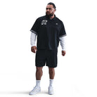 A bearded man stands confidently in a black HJ 2965010 Nike SB Dri-FIT Jacquard Skate Jersey over a white long-sleeve shirt, paired with black shorts, white crew socks, and white sneakers against a plain white background.