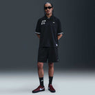 A man with braided hair, sunglasses, and tattoos stands against a dark background wearing the HJ 2965010 Black Nike SB Dri-FIT Jacquard Skate Jersey, black shorts, black Nike socks, and maroon Nike sneakers.