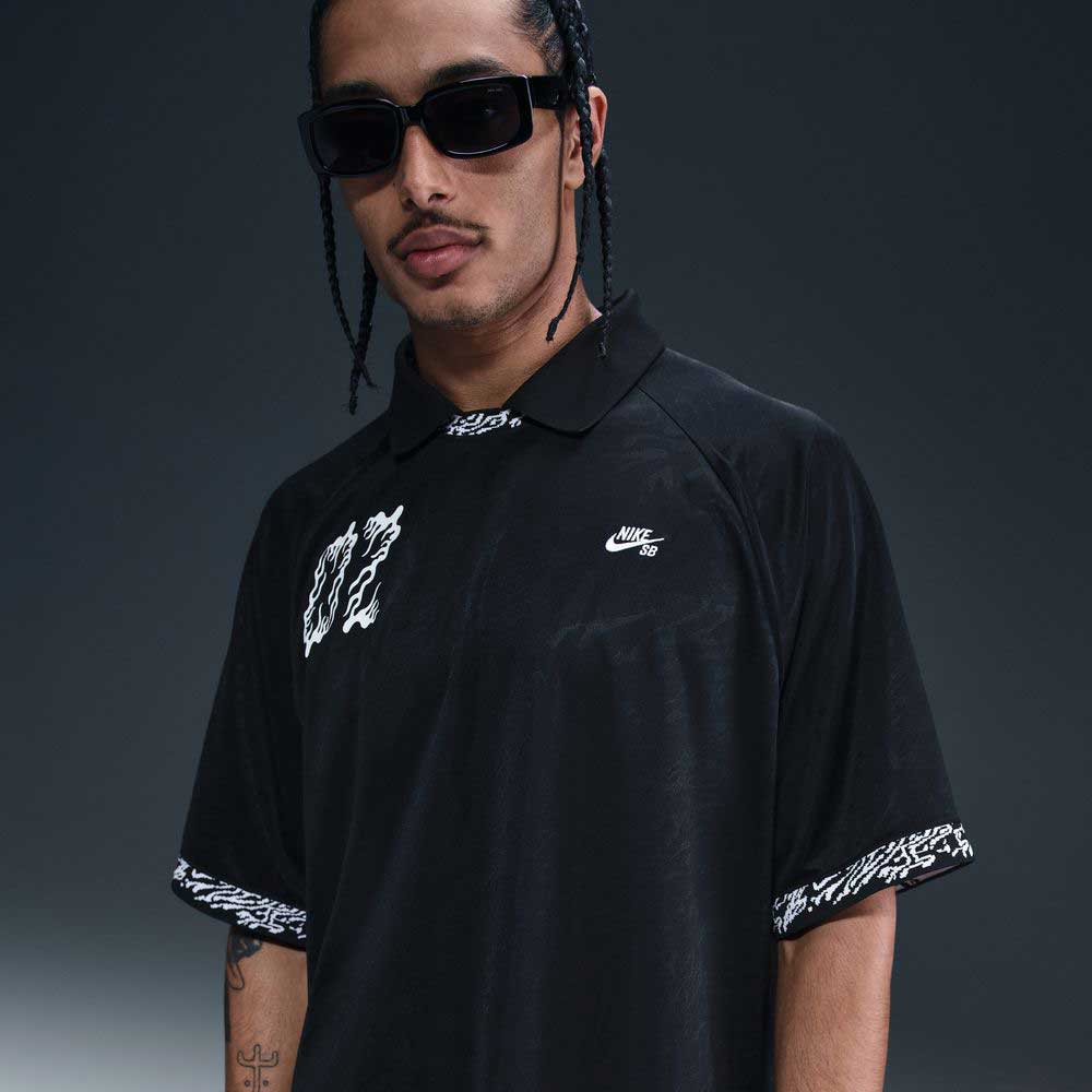 A man with braided hair and sunglasses wears the HJ 2965010 Black Nike SB Dri-FIT Jacquard Skate Jersey Skateboarding, featuring white chest and sleeve graphics, standing against a dark gradient background.