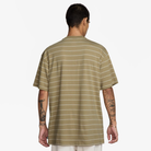 A person with short dark hair, visible arm tattoos, and light skin is seen from behind wearing the HJ0812297 U NK SB TEE M90 Tan White STRIPE Nike SB Skateboarding shirt and white pants against a plain background.