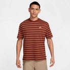 A young man stands facing forward, wearing the HJ0812619 U NK SB TEE M90 Maroon Red STRIPE by Nike SB Skateboarding and tan shorts, against a plain light background.