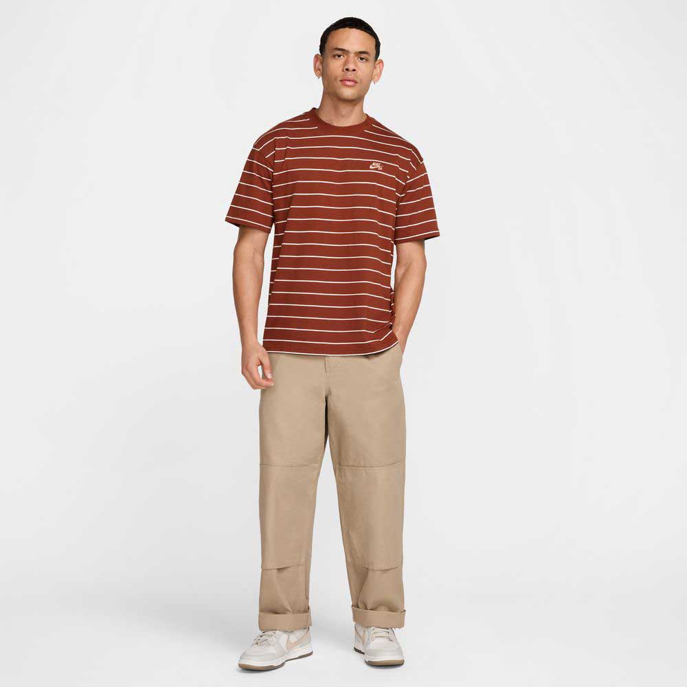 A man faces forward in a maroon red HJ0812619 U NK SB TEE M90 STRIPE Nike SB Skateboarding shirt, beige loose-fit pants, and white sneakers against a plain white background.