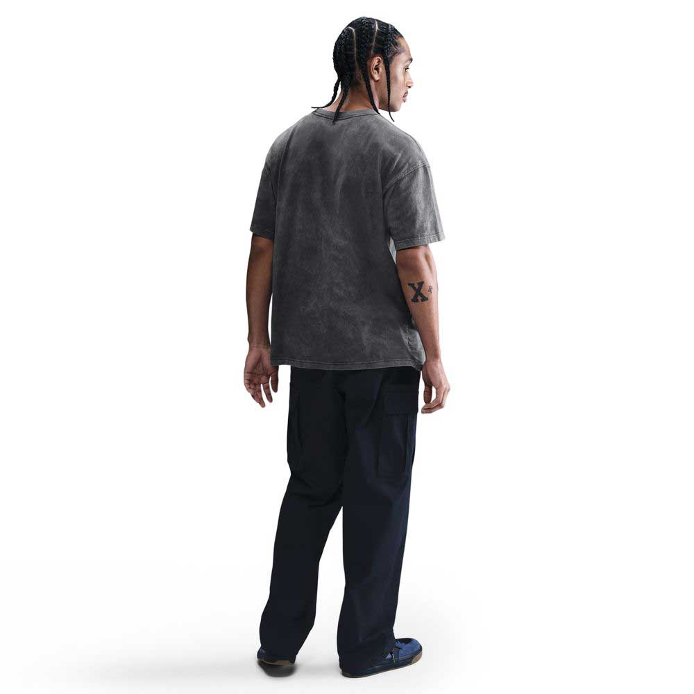 A person with braided hair, wearing the HJ0816060 Max 90 Gray Nike SB Skateboarding T-Shirt, stands with their back to the camera. An X tattoo is on their left forearm. The background is plain white.