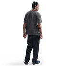 A person with braided hair, wearing the HJ0816060 Max 90 Gray Nike SB Skateboarding T-Shirt, stands with their back to the camera. An X tattoo is on their left forearm. The background is plain white.