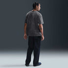 A person with braided hair faces away, wearing the Nike SB HJ0816060 Max 90 Skate T-Shirt in gray, dark cargo pants, and shoes. An X tattoo is visible on their left arm. The background is plain and dark.