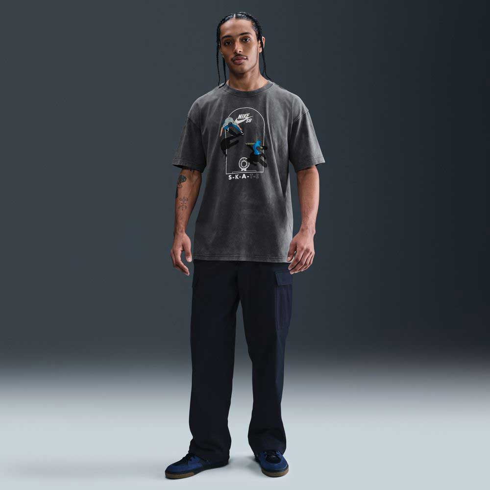 A man with braided hair stands facing forward in the Nike SB Max 90 Skate T-Shirt SB Tee Gray (HJ0816060) with screenprinted graphics, paired with dark cargo pants and sneakers, set against a plain dark background.