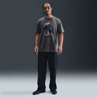 A man with braided hair stands facing forward in the Nike SB Max 90 Skate T-Shirt SB Tee Gray (HJ0816060) with screenprinted graphics, paired with dark cargo pants and sneakers, set against a plain dark background.