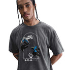 A person with braided hair wears the HJ0816060 Max 90 Skate T-Shirt— a gray Nike SB tee with a roomy fit, midweight cotton, and colorful skateboarding screenprinted graphics featuring "SKATE" on the front.