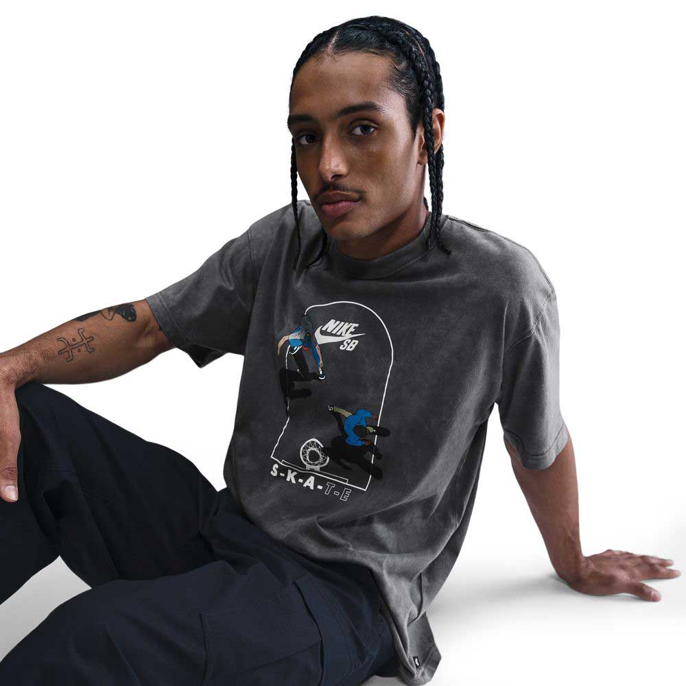 A person with braided hair sits on the floor wearing the gray HJ0816060 Max 90 Skate T-Shirt SB Tee by Nike SB, featuring a skateboarding screenprint design, paired with dark pants against a white background.