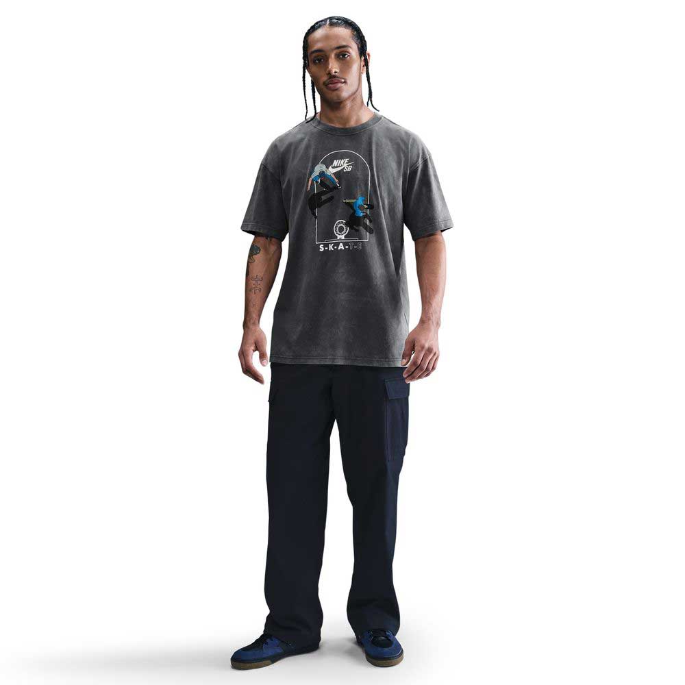 A man with braided hair stands facing forward in a gray Nike SB Max 90 Skate T-Shirt (HJ0816060) with a roomy fit, paired with black cargo pants and dark sneakers, set against a plain white background.