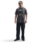 A man with braided hair stands facing forward in a gray Nike SB Max 90 Skate T-Shirt (HJ0816060) with a roomy fit, paired with black cargo pants and dark sneakers, set against a plain white background.