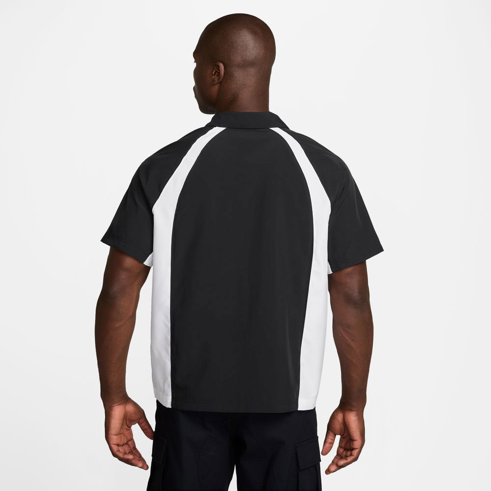 A man faces away from the camera, wearing a Nike SB HJ2956010 Black White Dri-Fit Woven Short-Sleeve Bowler Shirt with skateboarding embroidery and black pants, set against a plain white background.