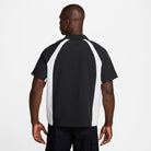 A man faces away from the camera, wearing a Nike SB HJ2956010 Black White Dri-Fit Woven Short-Sleeve Bowler Shirt with skateboarding embroidery and black pants, set against a plain white background.