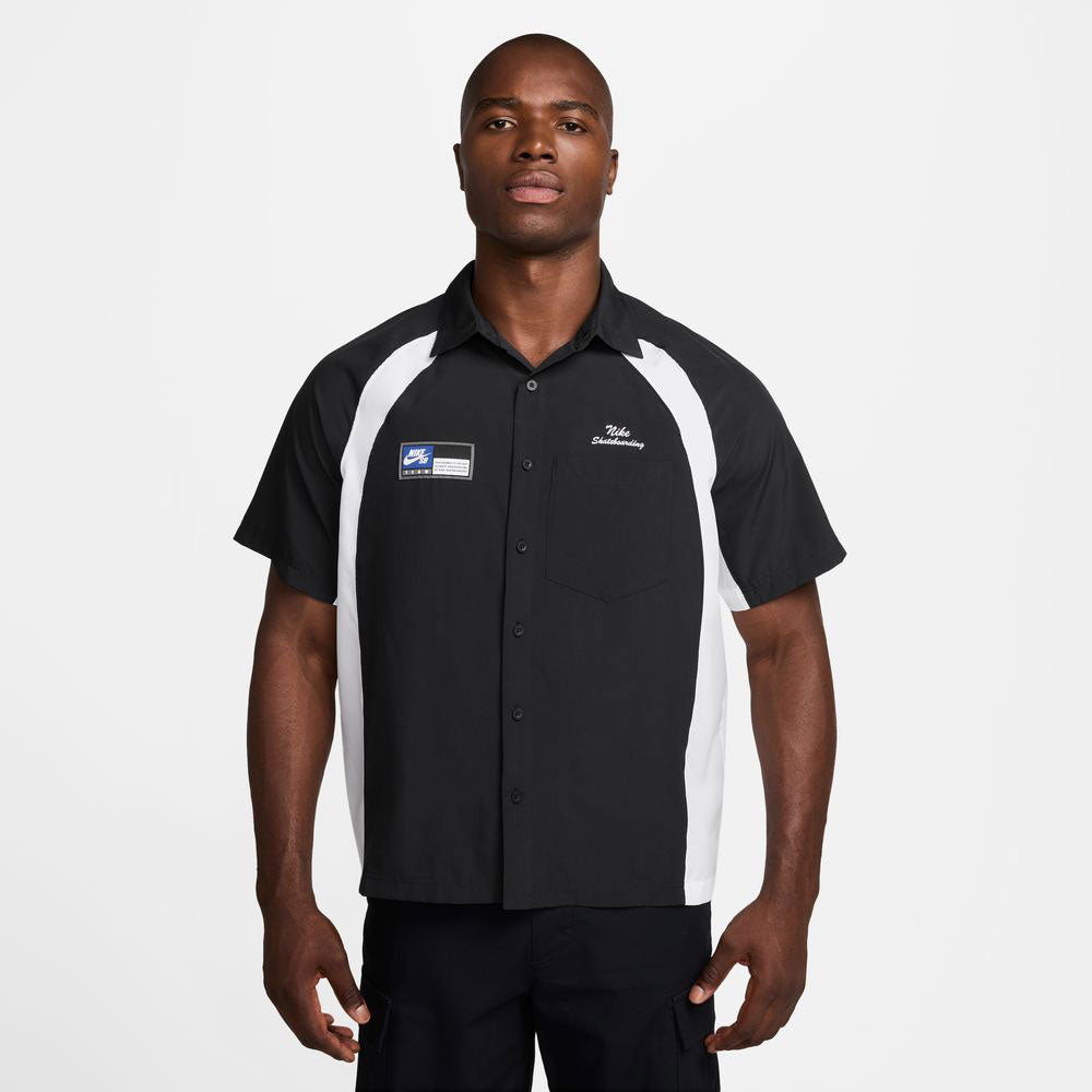 A man stands facing forward, wearing a HJ2956010 Black White Dri-Fit Woven Short-Sleeve Bowler Shirt by Nike SB Skateboarding, featuring embroidery and a chest pocket.