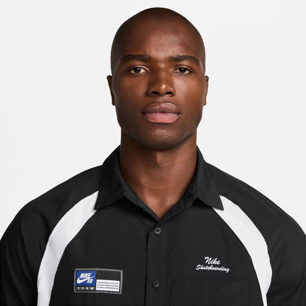 A man with short hair wears the HJ2956010 Black White Dri-Fit Woven Short-Sleeve Bowler Shirt by Nike SB, featuring skateboarding embroidery, standing against a plain white background.
