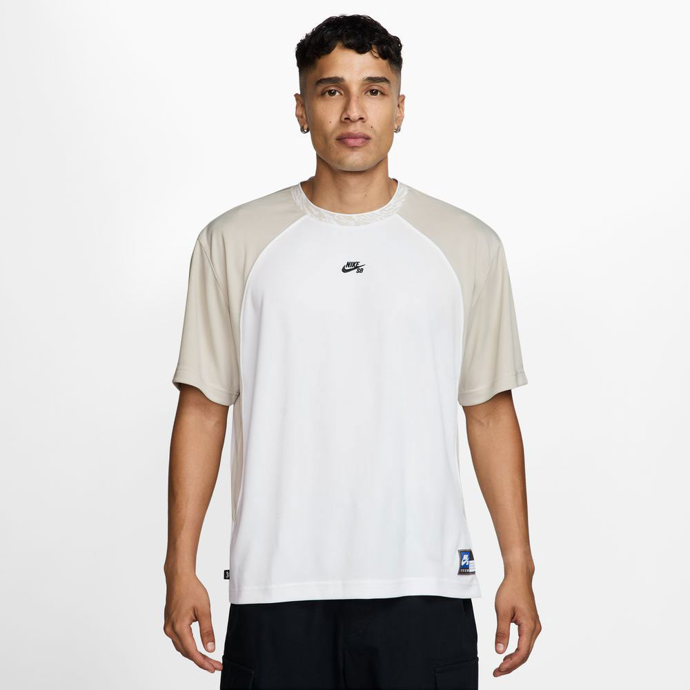 A man stands facing forward in the HJ2956010 Light Gray White Dri-Fit Knit Skate Jersey by Nike SB Skateboarding, featuring embroidered details, paired with black pants against a plain white background.