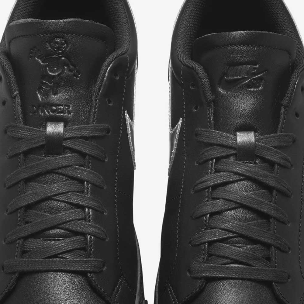 Close-up of two black HJ6703001 Black White Silver Blazer Low Dancer Nike SB leather skateboarding shoes. The left tongue has an embossed mascot and "DUNKER" while the right shows the classic Nike logo from the Dancer collaboration.