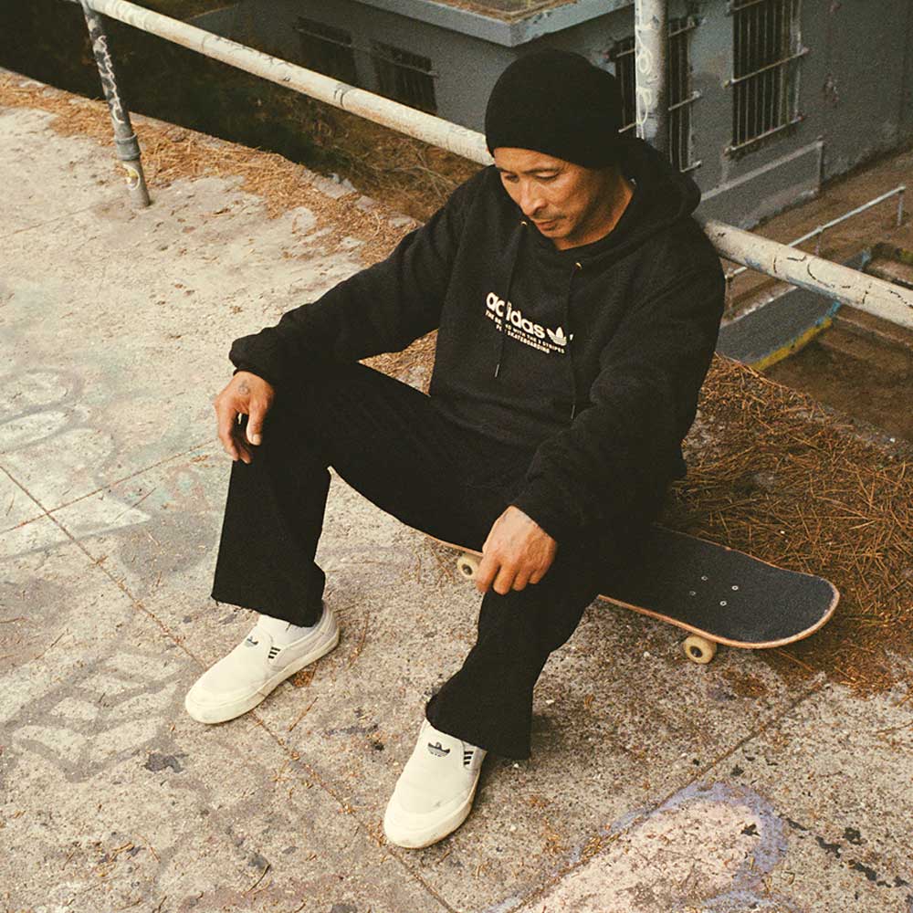 A person wearing black hoodie, black pants, and a black beanie sits on a skateboard in front of graffiti-covered railings, sporting HQ4721 Gray White Shmoofoil Slip RX Adidas Skateboarding Suede Shoes—ideal skate park footwear.