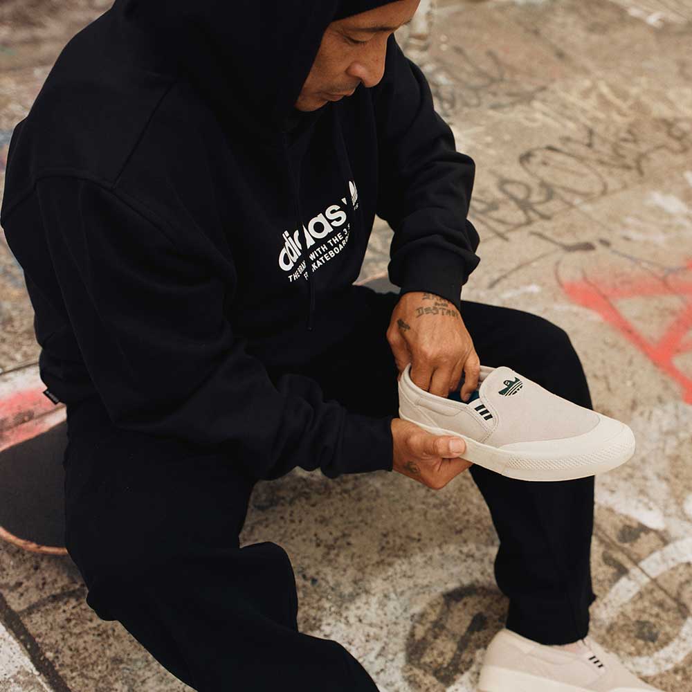 A person in a black hoodie and pants sits on graffiti-covered ground at a skate park, holding HQ4721 Gray White Shmoofoil Slip RX Adidas Skateboarding Suede Shoes, and appears to be putting them on.