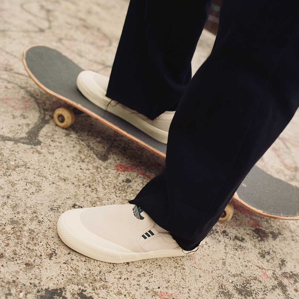 A person in black pants stands on a skateboard, wearing HQ4721 Gray White Shmoofoil Slip RX Adidas Skateboarding Suede Shoes—ideal footwear for testing tricks on graffiti-marked concrete.