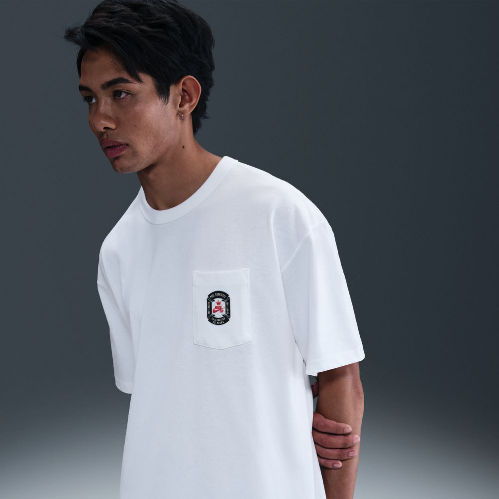 A young man with dark hair wears the Nike SB Prod 20 Year Tee HV1309100 in white, featuring a screenprinted pocket graphic. He stands against a dark gray background, looking to the side and highlighting daily comfort.