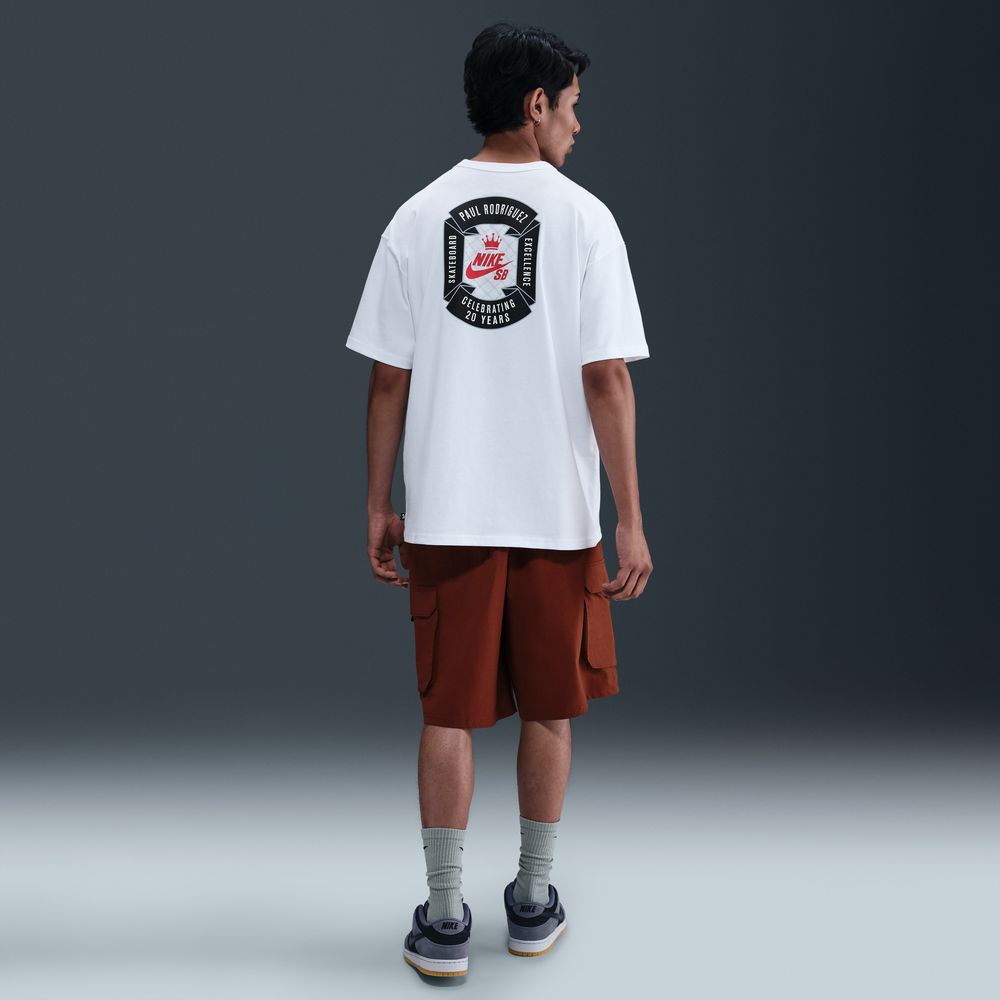 A man faces away, wearing the white Nike SB 20 Year Tee HV1309100 with screenprinted Max 90 Skate graphics, paired with brown cargo shorts, gray socks, and black sneakers against a plain dark background.