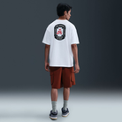 A man faces away, wearing the white Nike SB 20 Year Tee HV1309100 with screenprinted Max 90 Skate graphics, paired with brown cargo shorts, gray socks, and black sneakers against a plain dark background.