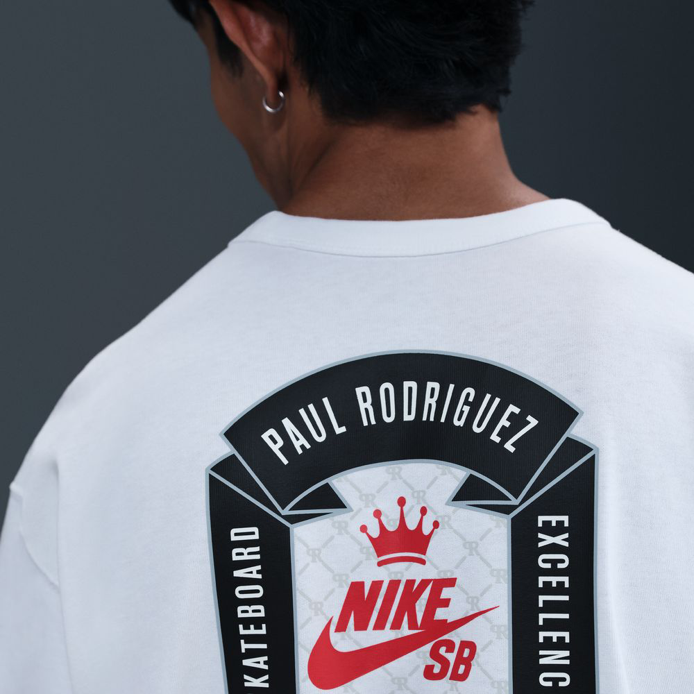 A person with short dark hair is seen from behind, wearing the Nike SB Prod 20 Year Tee HV1309100, a roomy white skate T-shirt featuring a screenprinted SB logo and Paul Rodriguez’s name with "Skateboard Excellence" printed around it.