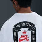 A person with short dark hair is seen from behind, wearing the Nike SB Prod 20 Year Tee HV1309100, a roomy white skate T-shirt featuring a screenprinted SB logo and Paul Rodriguez’s name with "Skateboard Excellence" printed around it.