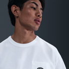 A person with short dark hair and a small hoop earring looks to the side, wearing the Nike SB Prod 20 Year Tee HV1309100 Max 90 Skate T-Shirt in white, featuring screenprinted details, against a plain dark background.
