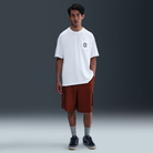 A man stands in a studio wearing the white Prod 20 Year Tee HV1309100 Max 90 Skate T-Shirt SB Tee by Nike SB with a small chest graphic, rust shorts, gray socks, and dark sneakers against a dark gray background.