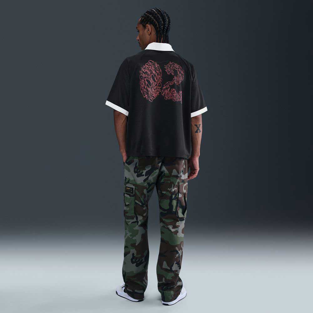 A person with braided hair faces away, wearing the Nike SB HV4457010 Polo Skate T-Shirt in black, red, and white with a screenprinted design, camo cargo pants, and white sneakers. The 100% cotton tee is machine washable.