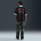 A person with braided hair faces away, wearing the Nike SB HV4457010 Polo Skate T-Shirt in black, red, and white with a screenprinted design, camo cargo pants, and white sneakers. The 100% cotton tee is machine washable.