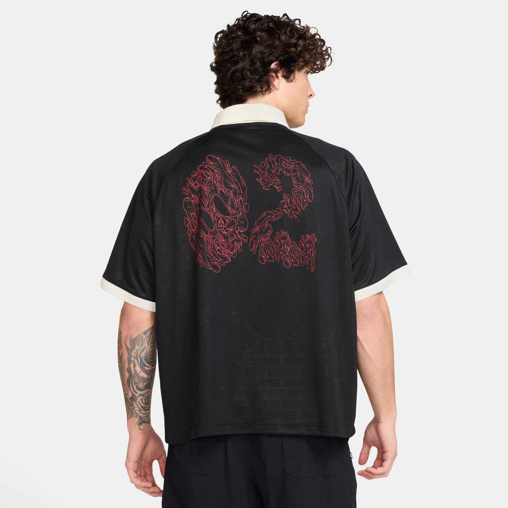 A man with curly hair, seen from the back, wears the Nike SB HV4457010 Polo Skate T-Shirt in black with white collar and cuffs, featuring a bold red "02" graphic on the back. Made of 100% cotton and machine washable for easy care.