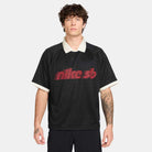 A young man with curly brown hair wears the HV4457010 Polo Skate T-Shirt SB Tee in black with a red and white Nike SB skateboarding logo, cream collar and cuffs. Made from 100% cotton, it's machine washable. He stands against a plain light gray background.