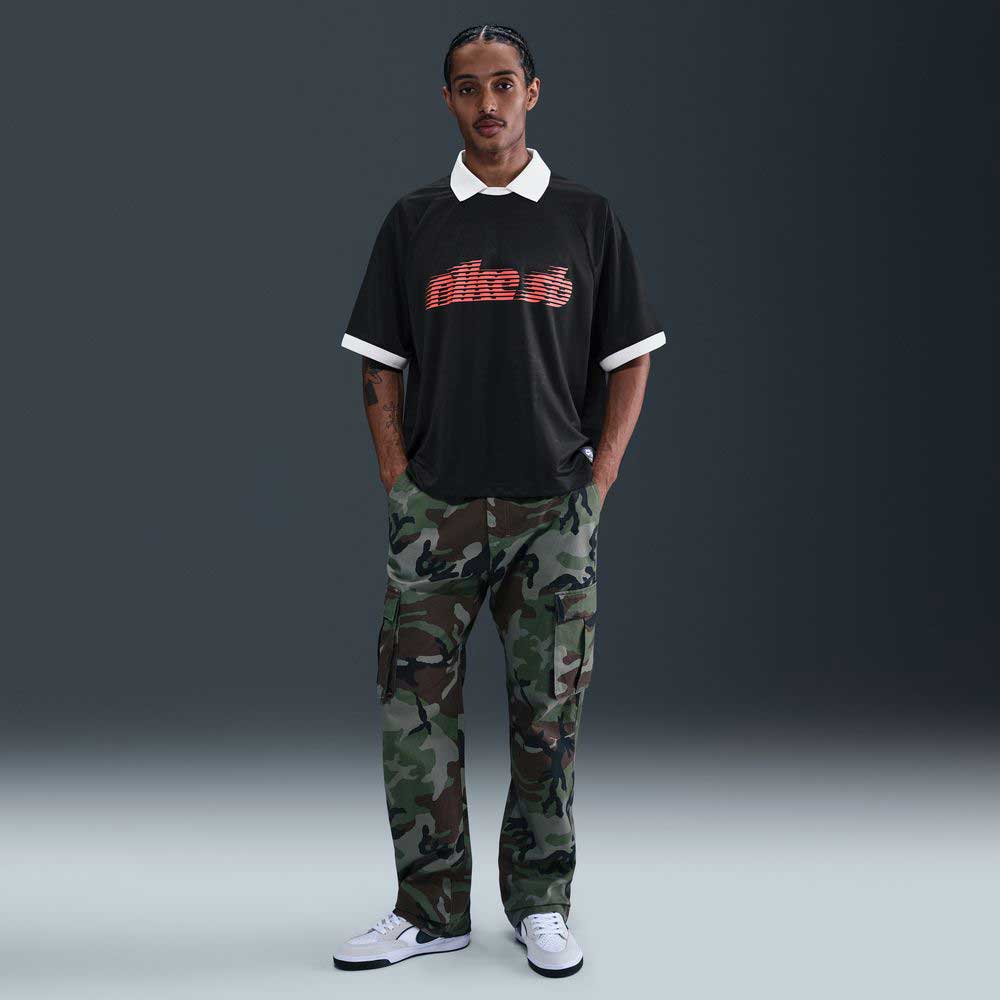 A man faces the camera in the HV4457010 Polo Skate T-Shirt SB Tee by Nike SB—a black, red, and white 100% cotton screenprinted skate shirt—paired with camo pants and white sneakers against a dark background. Machine washable.