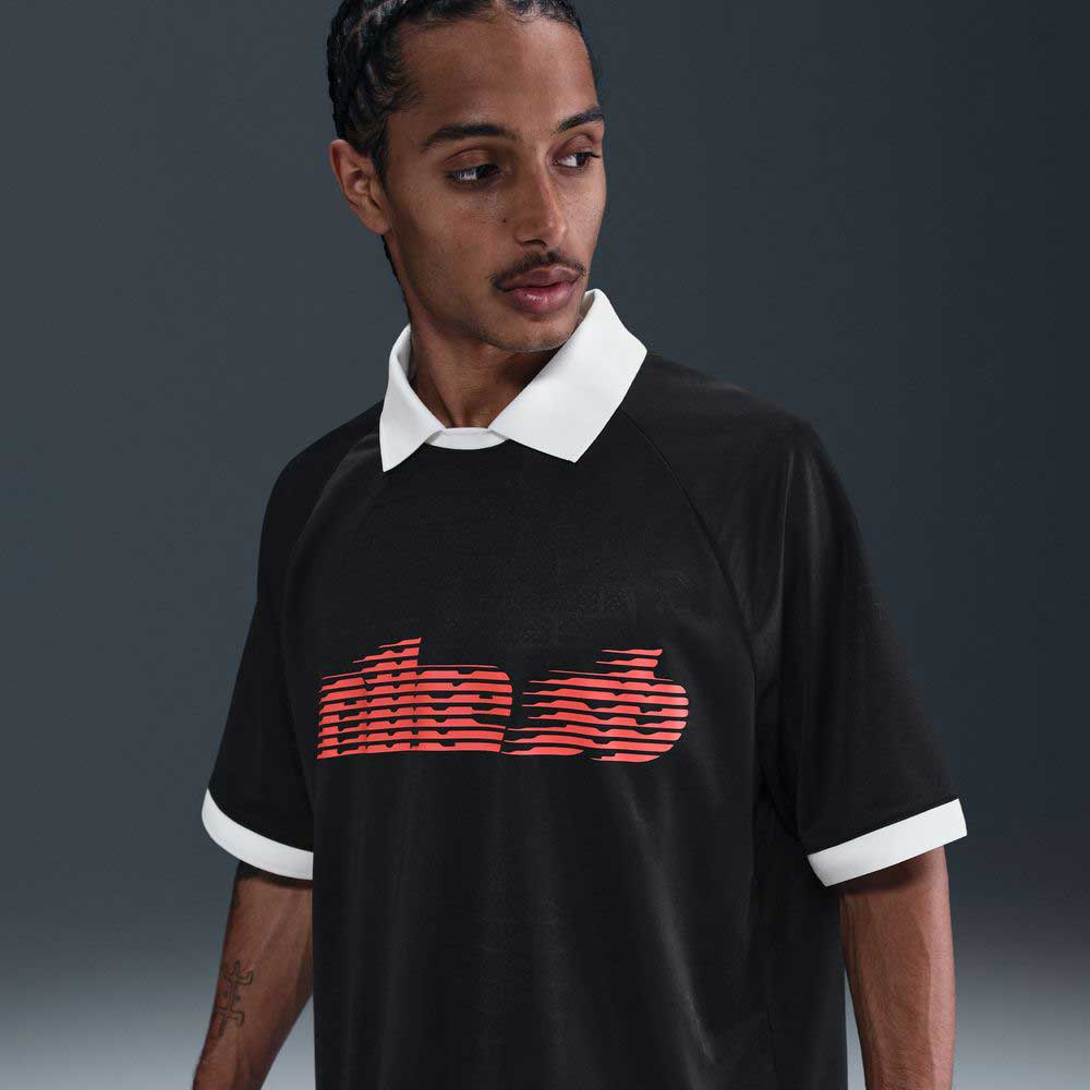 A man with braided hair wears the Nike SB HV4457010 Polo Skate T-Shirt, a black 100% cotton tee with white collar and cuffs and a red-orange striped screenprinted logo. He looks left, set against a dark blur. Machine wash recommended.