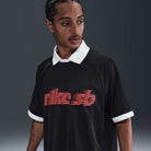 A man with braided hair wears the Nike SB HV4457010 Polo Skate T-Shirt, a black 100% cotton tee with white collar and cuffs and a red-orange striped screenprinted logo. He looks left, set against a dark blur. Machine wash recommended.