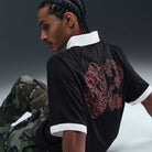 A man with braided hair sits on the floor in a HV4457010 Polo Skate T-Shirt SB Tee, black with a white collar and cuffs and an abstract red back design, paired with camo pants. The Nike SB Skateboarding shirt stands out against a gray gradient background.