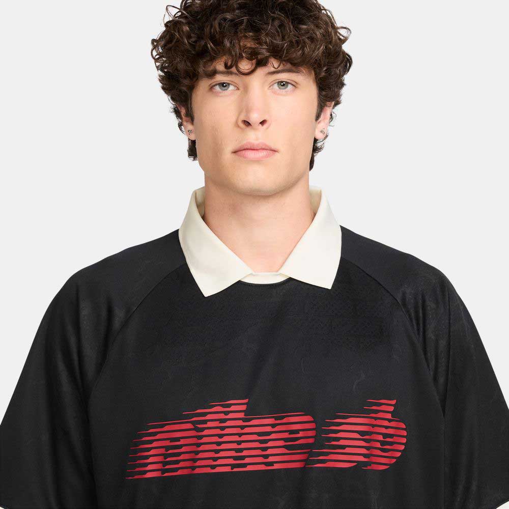 A person with curly brown hair wears the HV4457010 Polo Skate T-Shirt SB Tee in black with red and white Nike SB skateboarding screenprint, standing against a plain light background.
