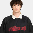 A person with curly brown hair wears the HV4457010 Polo Skate T-Shirt SB Tee in black with red and white Nike SB skateboarding screenprint, standing against a plain light background.