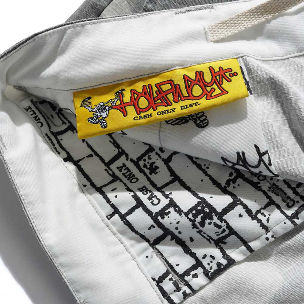 Close-up of the inside of the Halfway Shorts Grey Camouflage Ripstop Cash Only COD152601, showing a yellow HOLIDAY CASH ONLY DIST. label with cartoon graphics and a black-and-white text and brick patterned lining.